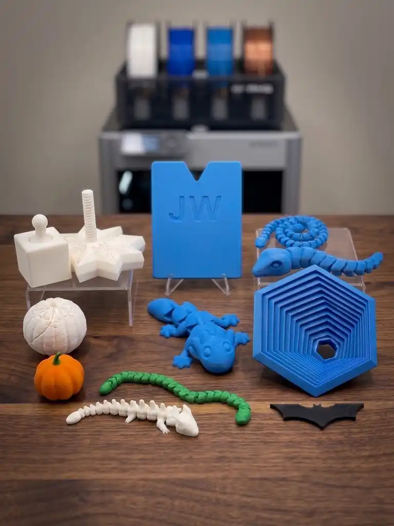 An assortment of 3D-printed pieces on a table in front of the printer, including dragons, coasters, a pumpkin and a nameplate